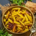 A brass bowl filled with spicy yellow Aloo Bhujia Sabji garnished with red chili, placed on burlap with naan bread, fresh cilantro, and a dried chili pepper in the background.