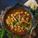 A bowl of Aloo Chole curry garnished with sliced green chilies and ginger, placed on a rustic cloth with naan bread, cilantro, and a spoon on the side.