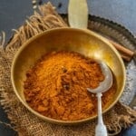 A brass bowl filled with bright orange pav bhaji masala powder and whole spices, with a metal spoon resting inside, placed on a burlap cloth and metal tray. Bay leaves and cinnamon sticks are visible in the background.