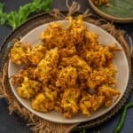A plate of crispy cabbage pakoda sits on a burlap cloth, accompanied by green chutney in a small bowl and a glass of chai. Fresh green chilies and coriander leaves are scattered around the setup.