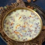 A brass bowl filled with creamy Doodh Pak dessert, garnished with saffron strands, small brown tapioca pearls, and placed on a rustic burlap mat with almonds and cardamom pods nearby.