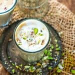 A glass of creamy Amritsari pista badam tea topped with sliced pistachios on a metal plate, with scattered pista and rustic burlap background.