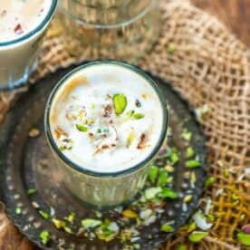 A glass of creamy Amritsari pista badam tea topped with sliced pistachios on a metal plate, with scattered pista and rustic burlap background.