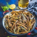 A bowl of crispy Kurkuri Bhindi garnished with spices, served with a slice of lemon and fresh coriander, with a glass of beverage and a napkin in the background.