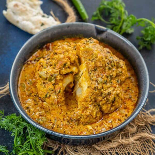 A bowl of Gobi Musallam features whole cauliflower covered in a creamy, orange-colored curry sauce, garnished with fresh cilantro, served with naan bread and green chili on the side.