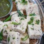 A plate of white, square pieces of steamed Oats Dhokla garnished with chopped cilantro, green chilies, mustard seeds, and curry leaves, served with tangy green chutney on the side.