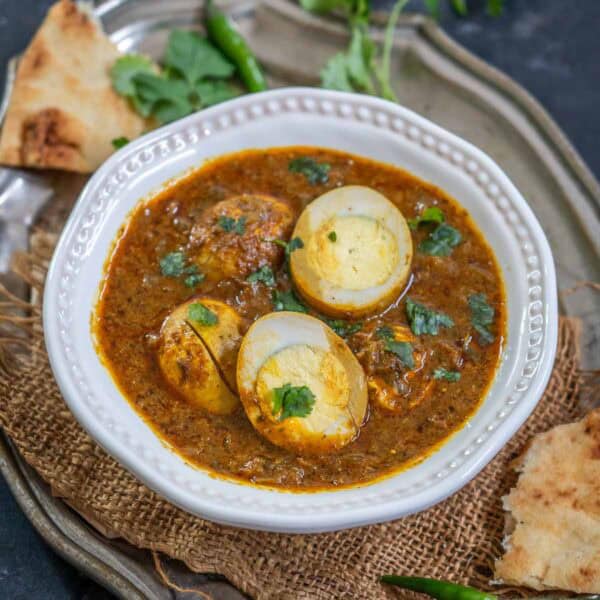 A white bowl of Dhaba Style Egg Curry garnished with cilantro, served with pieces of naan bread and green chilies on a rustic metal tray, set against a dark background.