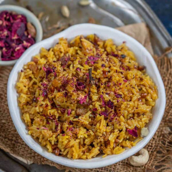 A bowl of Zarda, garnished with dried rose petals and spices, sits on a burlap mat atop a metal tray. Beside it, a small dish holds dried rose petals and whole cardamom pods.