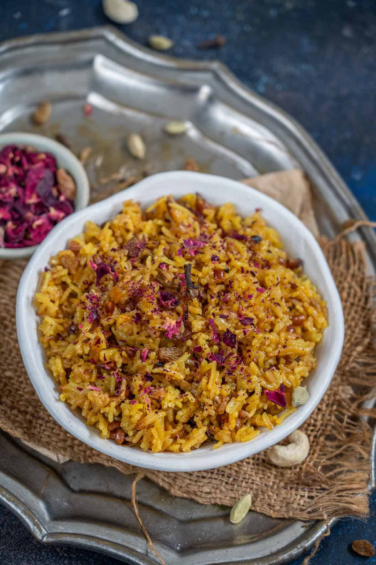 A bowl of Zarda, garnished with rose petals and nuts, is placed on a rustic metal tray with a small dish of dried rose petals beside it.