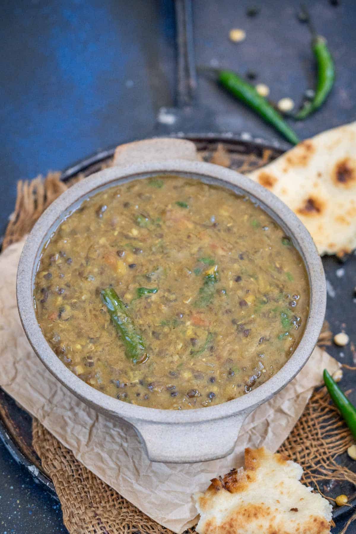 A bowl of Dal Amritsari with visible green chilies is placed on a piece of parchment paper, next to pieces of naan bread on a round tray.