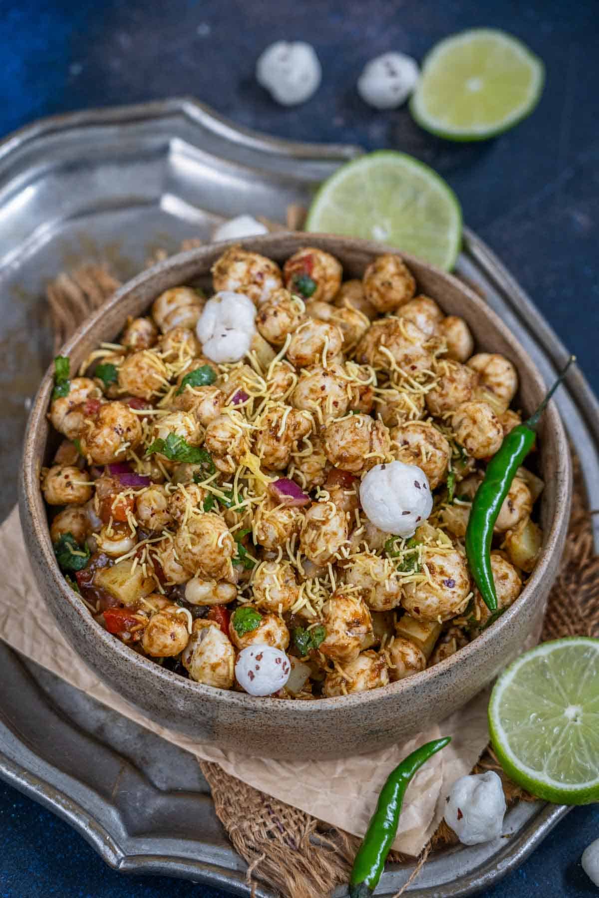 A bowl of Makhana Bhel, garnished with sev, chopped onions, coriander, and whole makhana, with green chilies and lime slices on the side, served on a rustic tray.