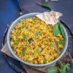 A bowl of Chana Pulao garnished with fresh cilantro and served alongside crispy papad pieces. Green chilies and mint leaves are scattered around on a dark surface.