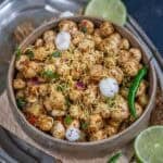 A bowl of Makhana Bhel, garnished with sev, chopped onions, coriander, and whole makhana, with green chilies and lime slices on the side, served on a rustic tray.