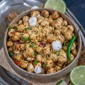 A bowl of Makhana Bhel, garnished with sev, chopped onions, coriander, and whole makhana, with green chilies and lime slices on the side, served on a rustic tray.