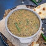 A bowl of Dal Amritsari with visible green chilies is placed on a piece of parchment paper, next to pieces of naan bread on a round tray.