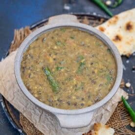 A bowl of Dal Amritsari with visible green chilies is placed on a piece of parchment paper, next to pieces of naan bread on a round tray.