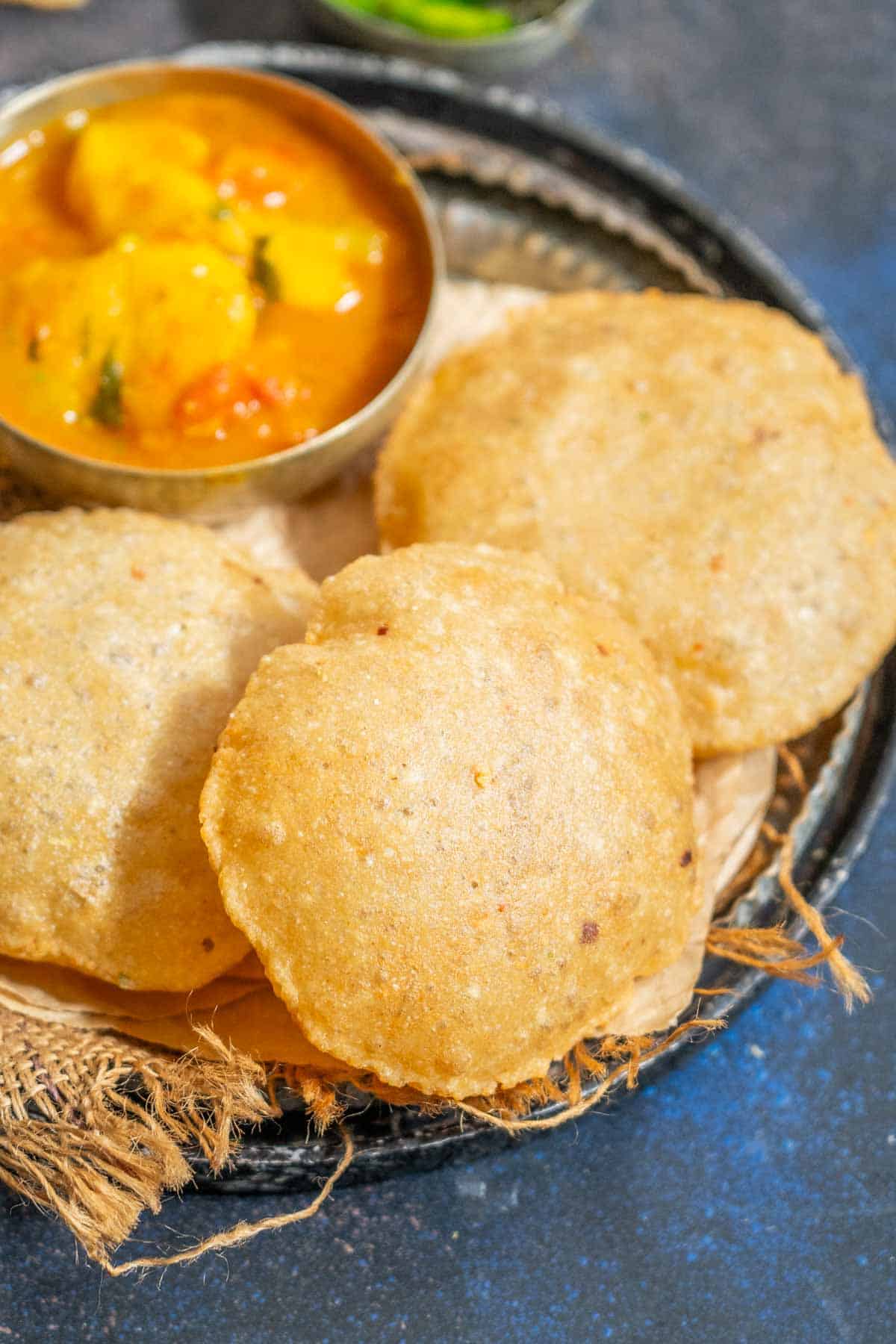 Three round, golden-brown crispy bedmi pooris are placed on a rustic tray lined with burlap, with a small bowl of curry visible in the top left corner.