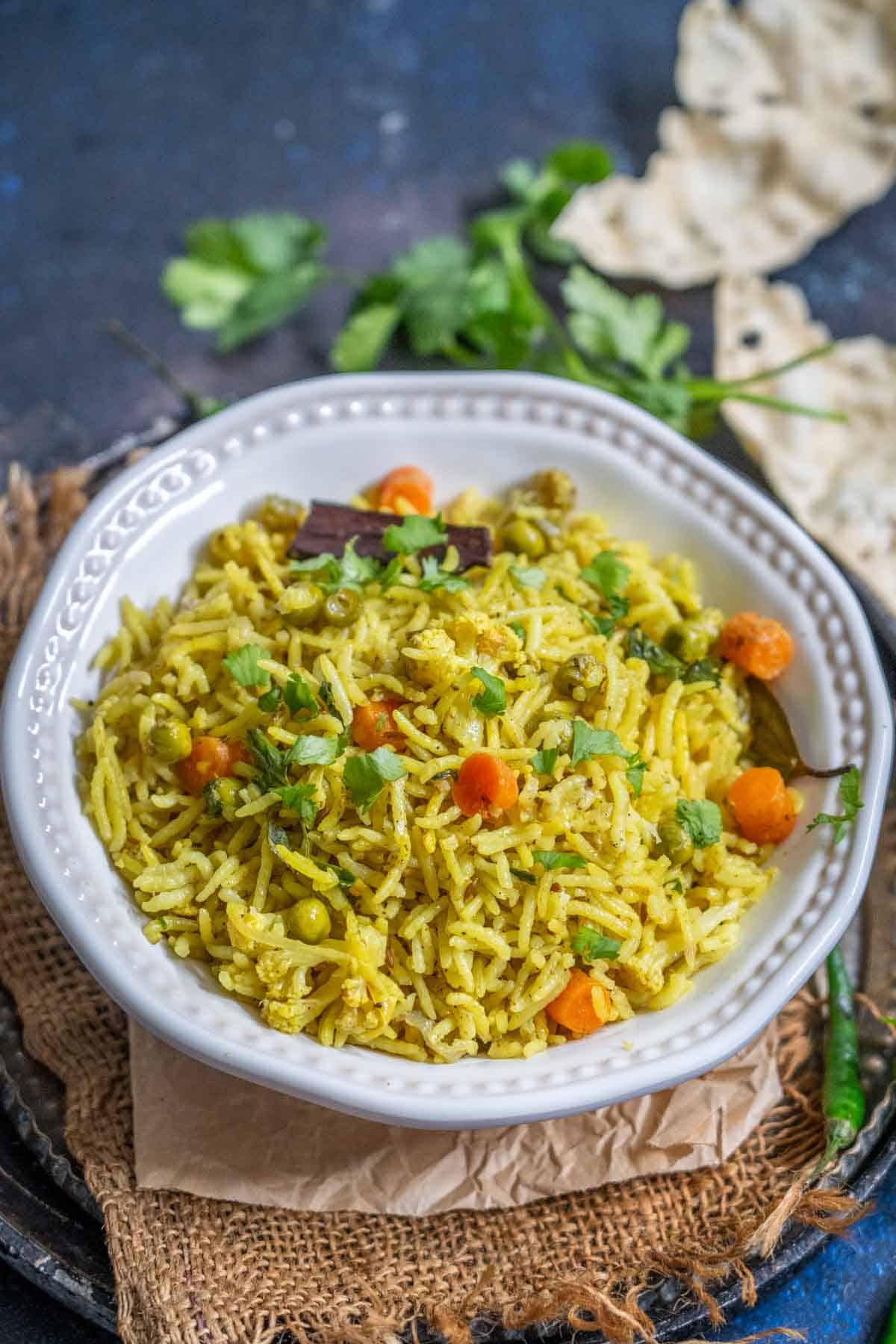 A white bowl filled with Chaas Wala Pulao, featuring yellow rice mixed with peas, carrots, and garnished with fresh cilantro. The bowl sits on a rustic woven mat.