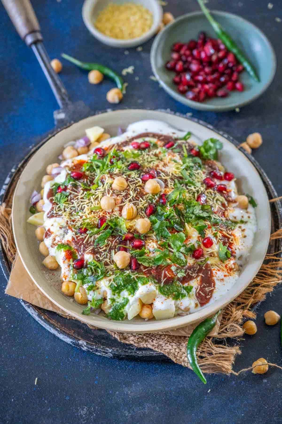 A plate of Dahi Chana chaat topped with yogurt, chickpeas, pomegranate seeds, green chutney, sev, fresh coriander, spices, and potato pieces on a rustic tray.