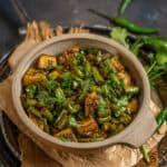 A ceramic bowl filled with North Indian Beans Aloo Sabzi, garnished with fresh cilantro, sits on a tray with green chilies and cilantro leaves in the background.