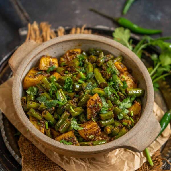 A ceramic bowl filled with North Indian Beans Aloo Sabzi, garnished with fresh cilantro, sits on a tray with green chilies and cilantro leaves in the background.