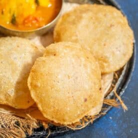 Three round, golden-brown crispy bedmi pooris are placed on a rustic tray lined with burlap, with a small bowl of curry visible in the top left corner.