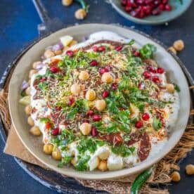 A plate of Dahi Chana chaat topped with yogurt, chickpeas, pomegranate seeds, green chutney, sev, fresh coriander, spices, and potato pieces on a rustic tray.