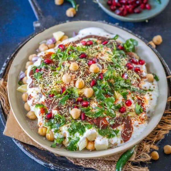 A plate of Dahi Chana chaat topped with yogurt, chickpeas, pomegranate seeds, green chutney, sev, fresh coriander, spices, and potato pieces on a rustic tray.