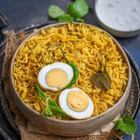 A bowl of yellow spiced Egg Pulao garnished with two halved boiled eggs, a green chili, mint leaves, and bay leaves, placed on a rustic cloth with a small bowl of raita in the background.