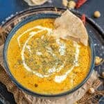 A bowl of creamy, orange-colored Malai Chole curry is garnished with a swirl of cream and herbs, with a piece of crispy poori resting on the rim. The bowl sits on a rustic tray with scattered chickpeas nearby.