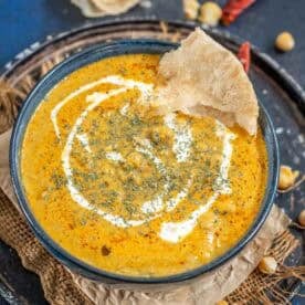 A bowl of creamy, orange-colored Malai Chole curry is garnished with a swirl of cream and herbs, with a piece of crispy poori resting on the rim. The bowl sits on a rustic tray with scattered chickpeas nearby.