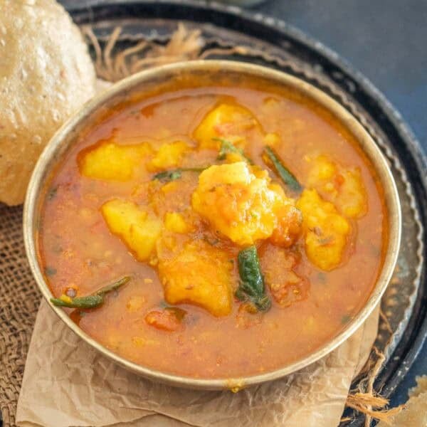 A bowl of North Indian Aloo Tamatar Ki Sabji, featuring yellow potato curry with green chili peppers in a rich, orange-red gravy, served on a dark tray with a side of puffed bread.