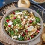 A bowl of churmur chaat, an Indian street food snack, is garnished with chopped cilantro, pomegranate seeds, and crushed puri, and placed on a tray with extra puri and spices in the background.