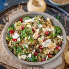 A bowl of churmur chaat, an Indian street food snack, is garnished with chopped cilantro, pomegranate seeds, and crushed puri, and placed on a tray with extra puri and spices in the background.