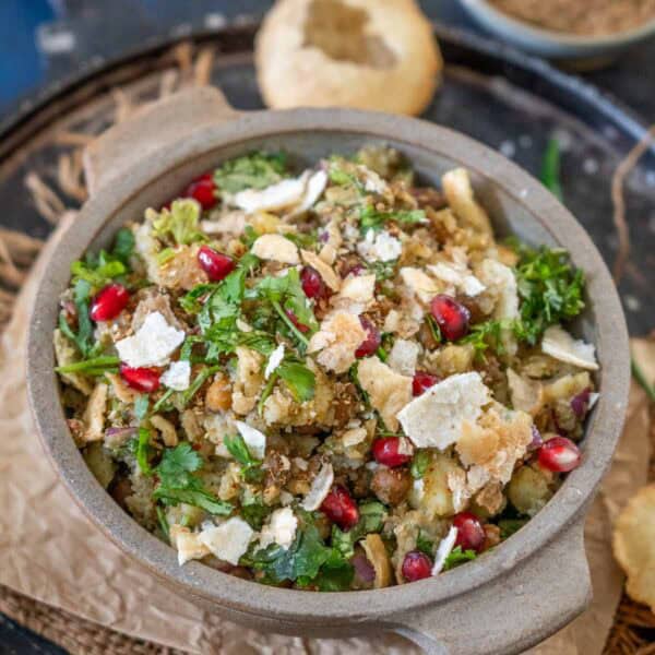 A bowl of churmur chaat, an Indian street food snack, is garnished with chopped cilantro, pomegranate seeds, and crushed puri, and placed on a tray with extra puri and spices in the background.