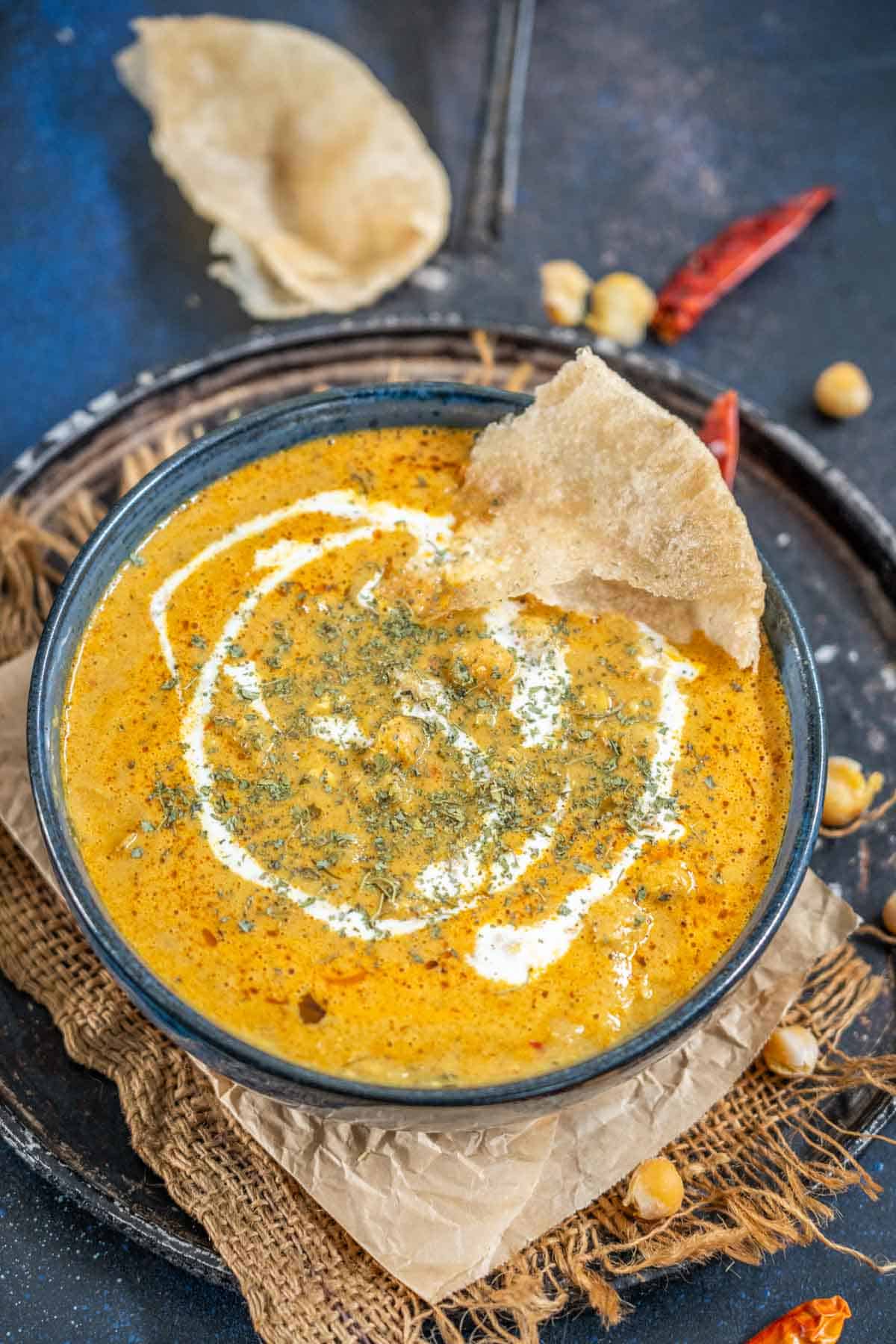 A bowl of creamy, orange-colored Malai Chole curry is garnished with a swirl of cream and herbs, with a piece of crispy poori resting on the rim. The bowl sits on a rustic tray with scattered chickpeas nearby.