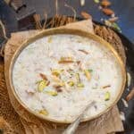 A bowl of creamy Doodh Dalia garnished with sliced almonds and pistachios. A spoon rests inside, and the bowl sits on brown paper atop rustic fabric.