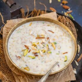 A bowl of creamy Doodh Dalia garnished with sliced almonds and pistachios. A spoon rests inside, and the bowl sits on brown paper atop rustic fabric.