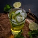 A glass of iced Saffron Nimbu Pani garnished with mint leaves and saffron strands, placed on a brown cloth with lime slices and mint in the background.