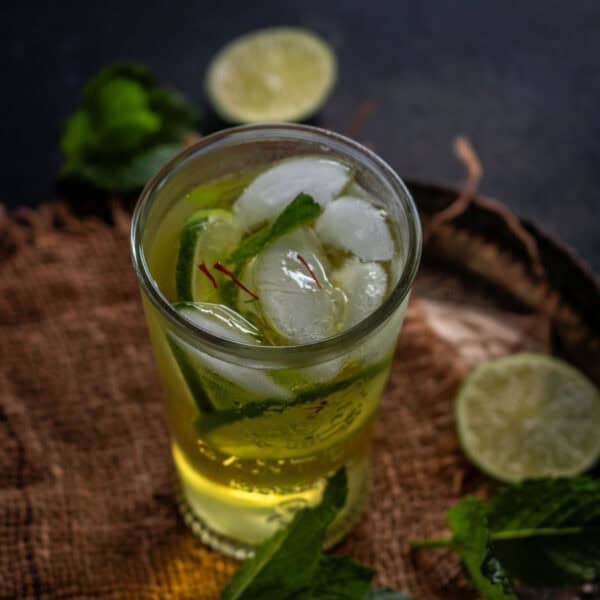 A glass of iced Saffron Nimbu Pani garnished with mint leaves and saffron strands, placed on a brown cloth with lime slices and mint in the background.