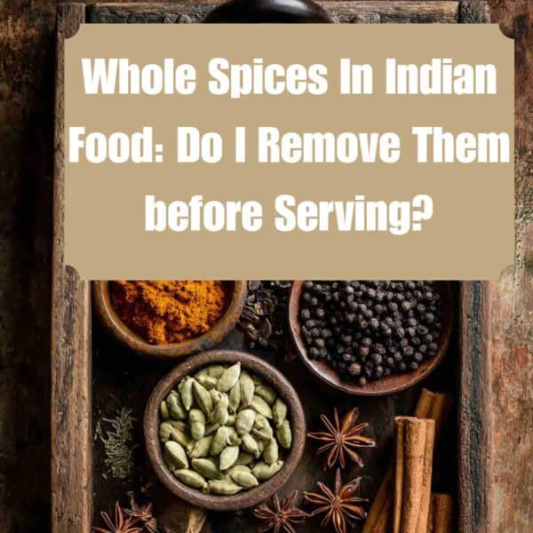 A rustic wooden tray holds bowls of whole spices such as peppercorns, star anise, cinnamon sticks, and more. Text overlay asks if you should remove whole spices before serving Indian food.