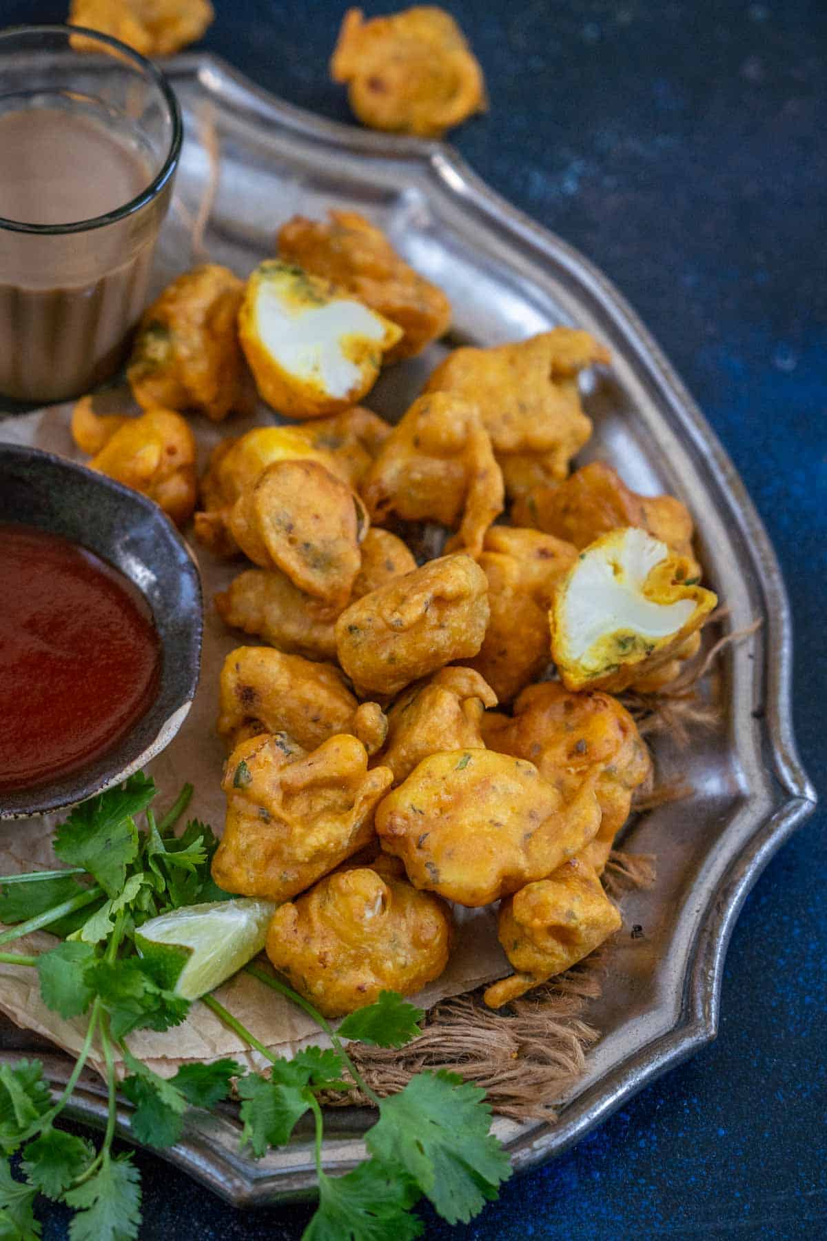 A plate of golden-brown gobi pakora is served with a bowl of red dipping sauce and fresh cilantro on the side. Some fritters are whole, while one is broken open, revealing the white cauliflower inside.
