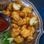 A plate of golden-brown gobi pakora is served with a bowl of red dipping sauce and fresh cilantro on the side. Some fritters are whole, while one is broken open, revealing the white cauliflower inside.