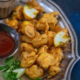 A plate of golden-brown gobi pakora is served with a bowl of red dipping sauce and fresh cilantro on the side. Some fritters are whole, while one is broken open, revealing the white cauliflower inside.