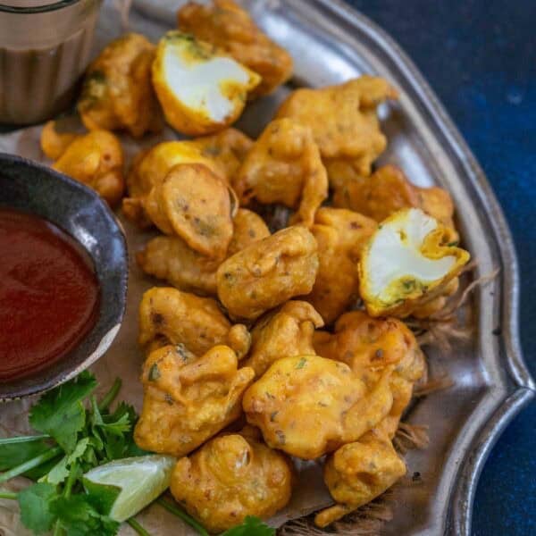 A plate of golden-brown gobi pakora is served with a bowl of red dipping sauce and fresh cilantro on the side. Some fritters are whole, while one is broken open, revealing the white cauliflower inside.