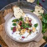 A bowl of Dahi Poha garnished with peanuts, pomegranate seeds, cilantro, and spices, with a crisp papad dipped in. The bowl sits on crumpled paper, surrounded by fresh cilantro and more papad.
