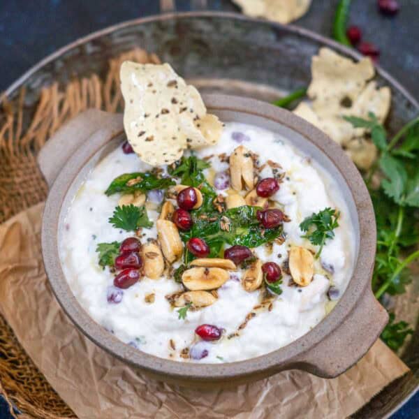 A bowl of Dahi Poha garnished with peanuts, pomegranate seeds, cilantro, and spices, with a crisp papad dipped in. The bowl sits on crumpled paper, surrounded by fresh cilantro and more papad.