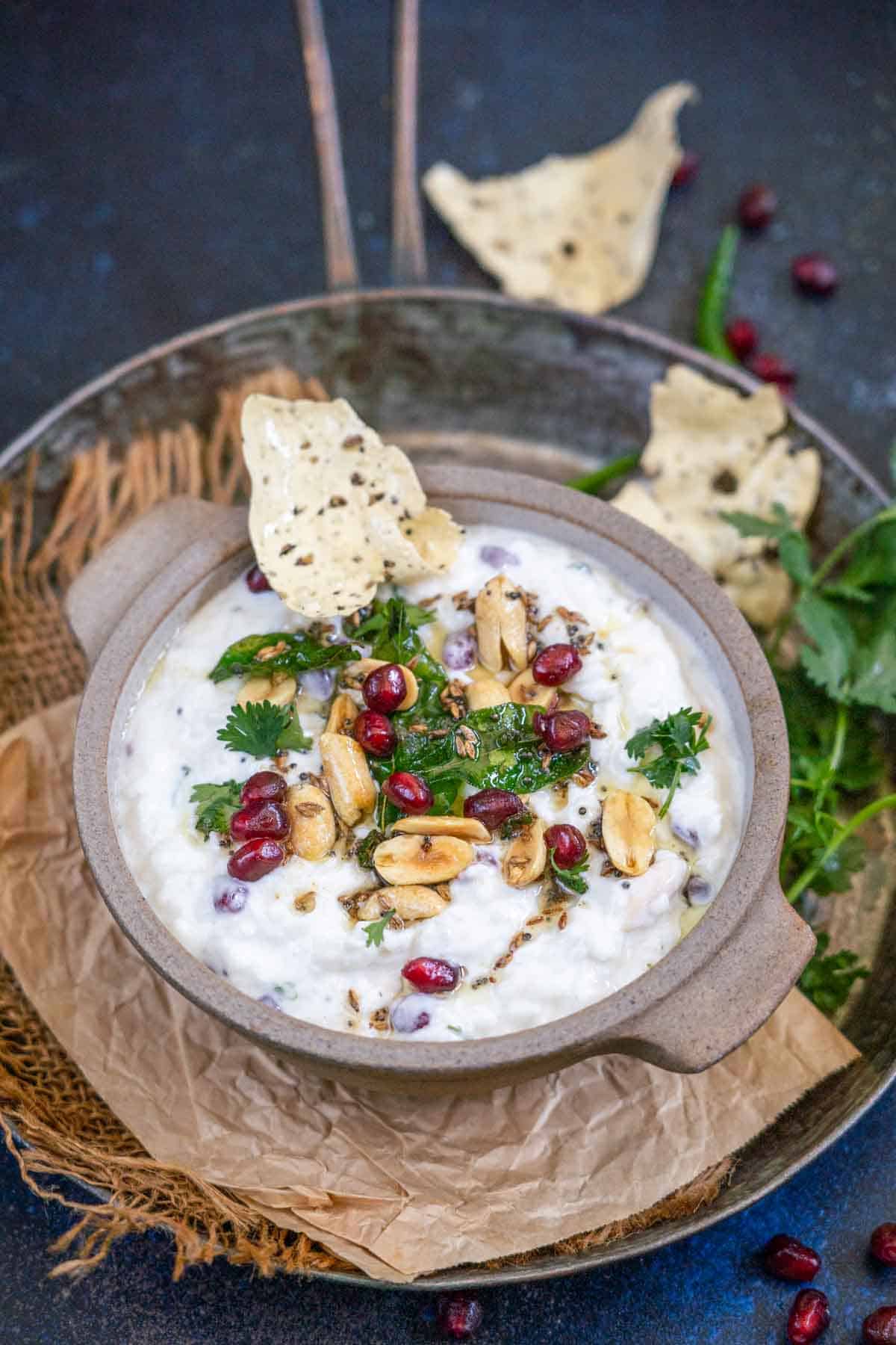 A bowl of Dahi Poha garnished with peanuts, pomegranate seeds, cilantro, and spices, with a crisp papad dipped in. The bowl sits on crumpled paper, surrounded by fresh cilantro and more papad.