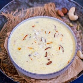 A bowl of creamy Samak Rice Kheer, also known as Barnyard Millet Kheer, garnished with chopped nuts and saffron strands sits on a rustic surface, with cashews and cardamom nearby—inviting you to try this delightful recipe.