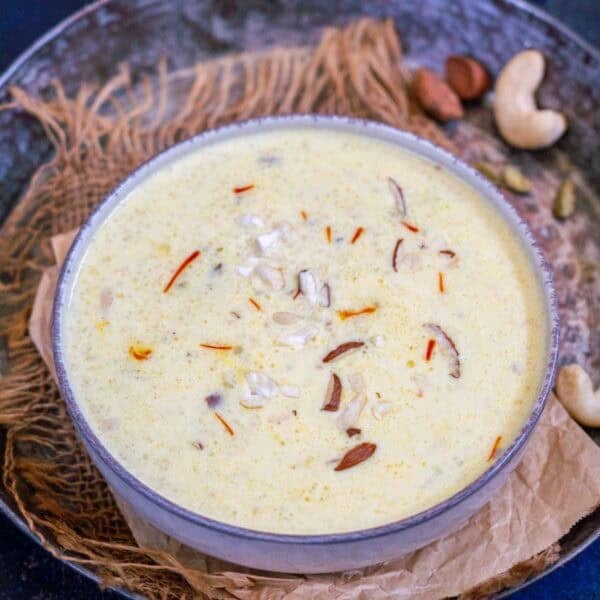 A bowl of creamy Samak Rice Kheer, also known as Barnyard Millet Kheer, garnished with chopped nuts and saffron strands sits on a rustic surface, with cashews and cardamom nearby—inviting you to try this delightful recipe.